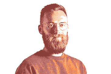 Close-up portrait of a bearded Thomas in his thirties, wearing round glasses and an orange sweater.